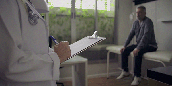 Closeup of female doctor holding clipboard and pen as middle-aged male patient waits in exam room background