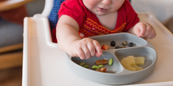 a young toddler eating a meal from a divided silicone plate (fruit, veggies, soy beans)